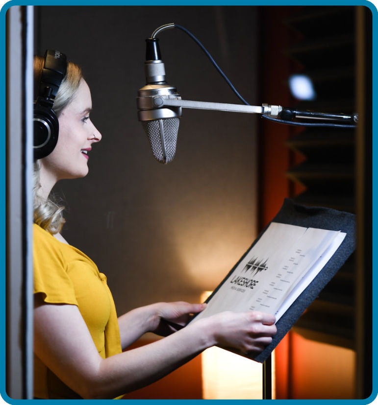 A narrator, a woman with shoulder length blonde hair, wears black headphones and a yellow top as she narrates a described video script for Lakeshore Media Services. The narrator stands in a warmly lit recording booth and speaks into a microphone.